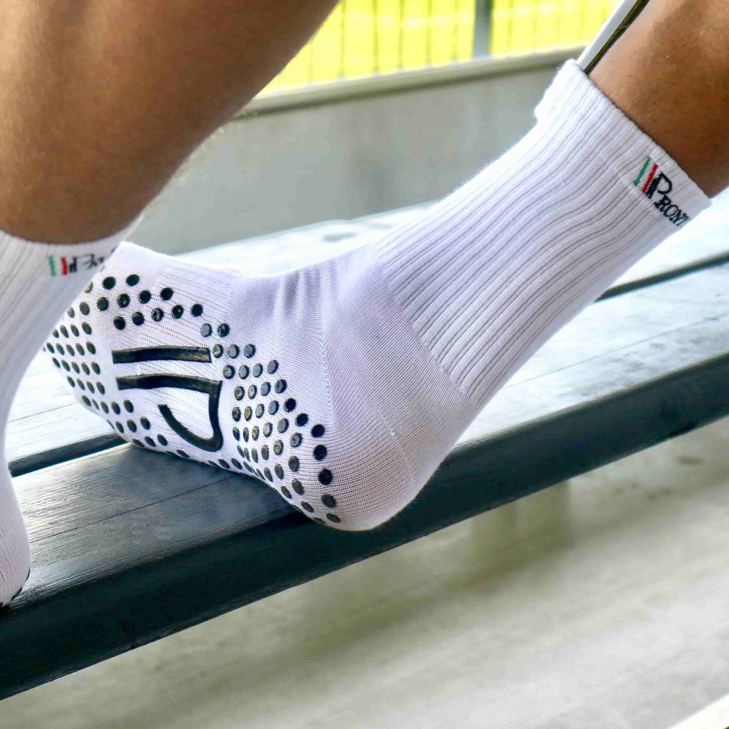 Et par fødder iført hvide Pronto gripsokker hviler på en træbænk med et stadion i baggrunden. Den ene sok viser det italiensk-inspirerede logo på anklen, mens den anden fremhæver de sorte grebpuder og "P"-logoet under sålen, designet til maksimal stabilitet og kontrol på banen.