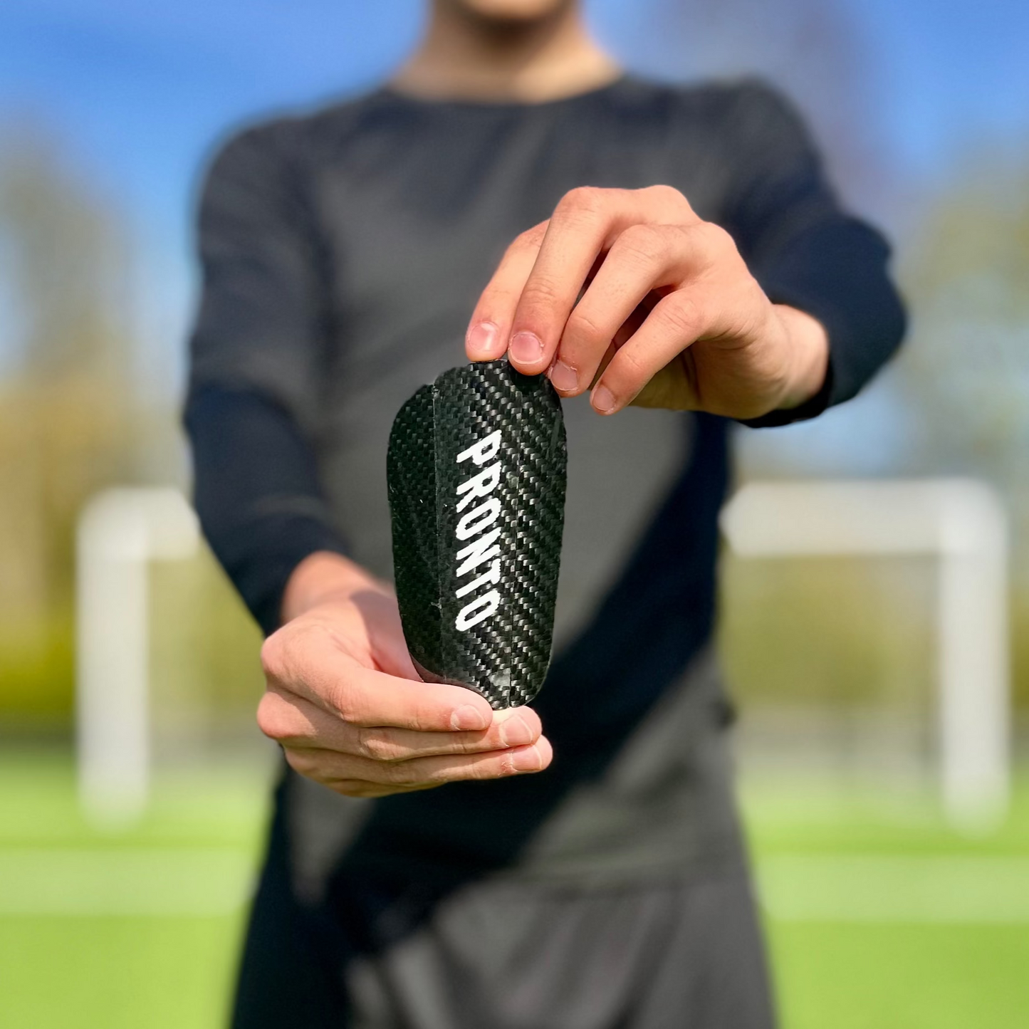En person holder en carbon letvægtsshinguard med 'PRONTO' trykt på den, med fodboldmål i baggrunden.