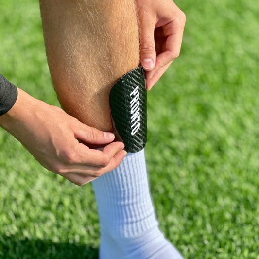 Hænder justerer en sort fodboldskinne med teksten "PRONTO" på skinnebenet, iført hvide gripsokker, på grøn kunstgræsbane.