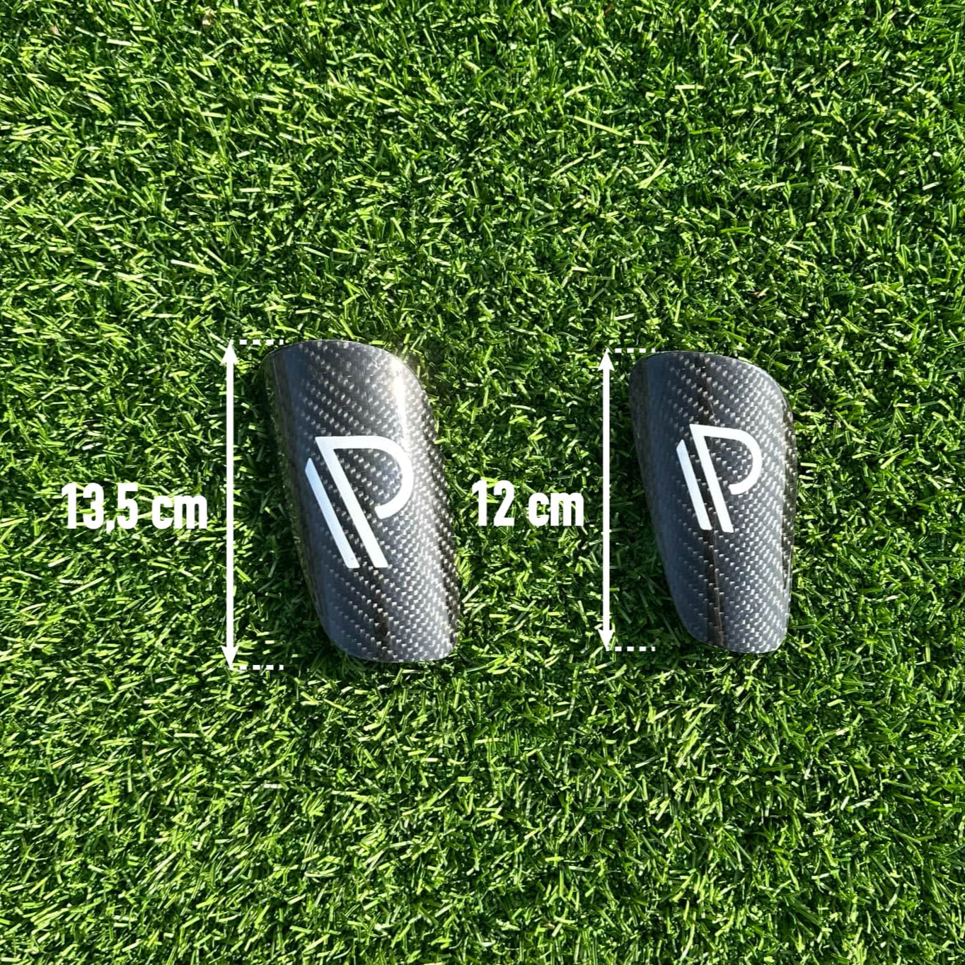 To sorte fodboldskinner med minimalistisk logo på grøn kunstgræsbane, målt til henholdsvis 13,5 cm og 12 cm i længde.