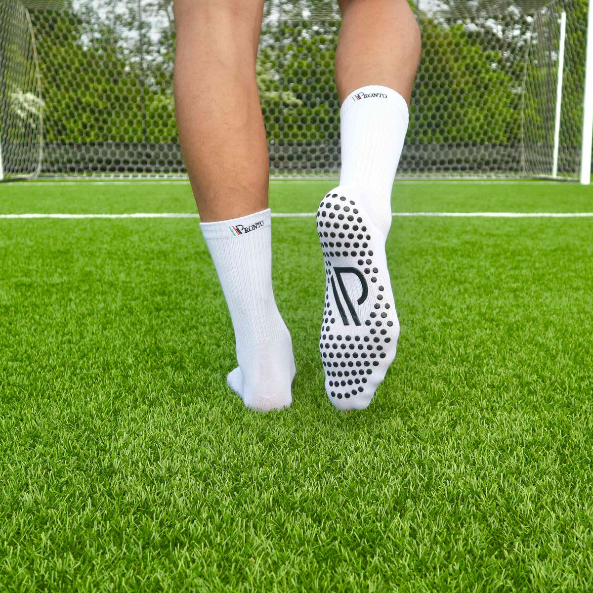 En fodboldspiller går væk fra kameraet på en grøn bane med et mål i baggrunden. Han har hvide Pronto gripsokker på, der viser det sorte grebmønster og logoet "P" under sålen for maksimal komfort og stabilitet.