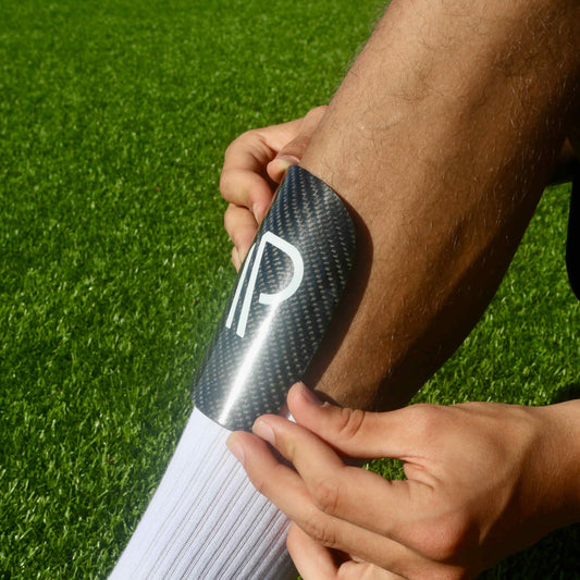 Hænder placerer en sort fodboldskinne med minimalistisk logo på skinnebenet, med grøn kunstgræsbane i baggrunden