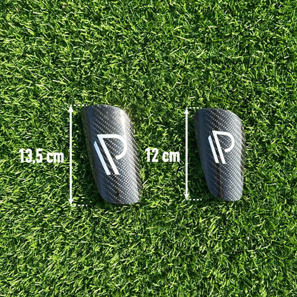 To sorte fodboldskinner med minimalistisk logo på grøn kunstgræsbane, målt til henholdsvis 13,5 cm og 12 cm i længde.